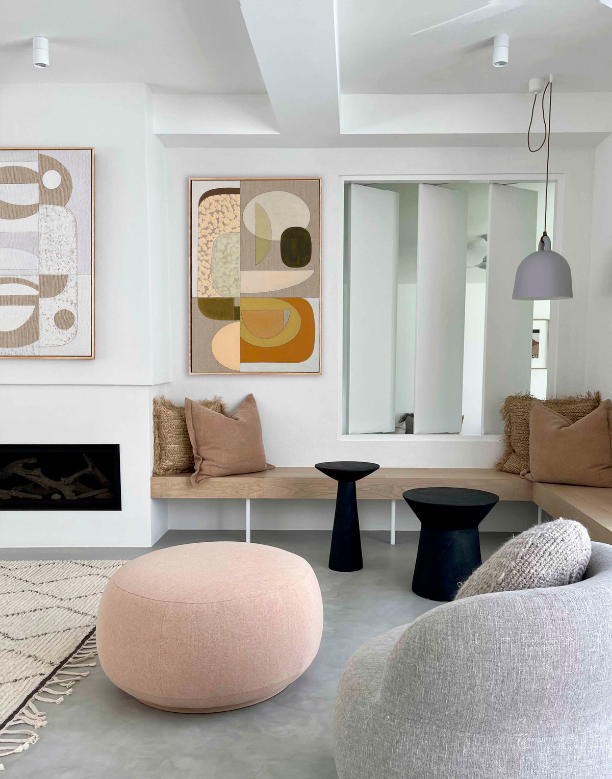 A modern living room interior featuring abstract wall art in beige, brown, and ochre tones, a light wood bench with tan pillows, a round blush pink pouf, black side tables, and a gray upholstered chair in a bright, contemporary space.