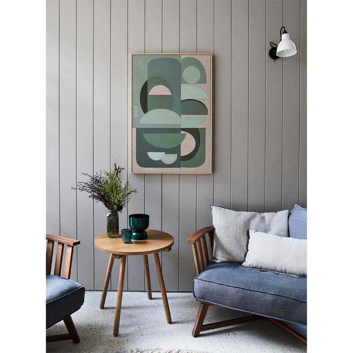 Framed wall art print featuring abstract geometric shapes in green, displayed on a wall above a wooden table with a plant and a lamp.