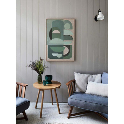 Framed wall art print featuring abstract geometric shapes in green, displayed on a wall above a wooden table with a plant and a lamp.